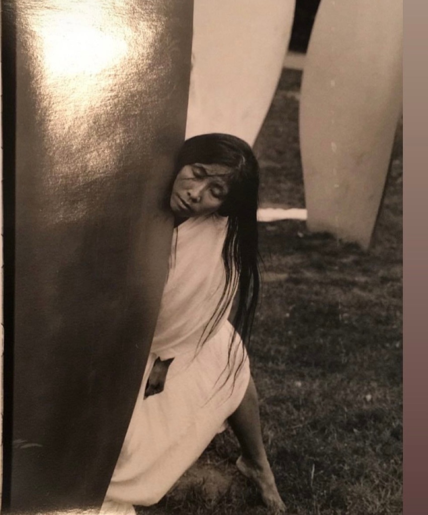 The artist Wannappa P-Eubanks leans with her face and full body against the smooth metal petal from Yoko Ono's sculpture, Skylanding. She wears a white flowing fabric and her long dark hair drapes across her shoulder.