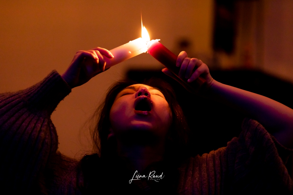 Artist Jinlu Luo holding two burning candles over her open mouth, her head is back with eyes closed. One is red and one is white, the light is orange and dark.