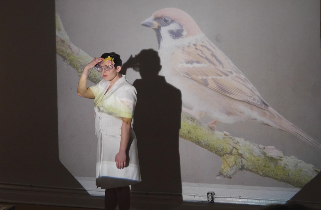 Artist Aurora Tabar dressed in a white lab coat stands in front of a projection of a songbird. She has a garland of flowers on her temple and holds her hand to her forehead, creating an angle with her elbow.