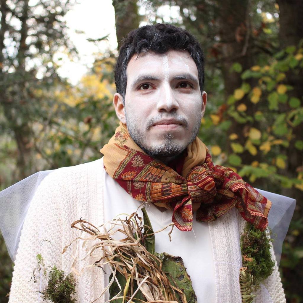 A portrait of artist Ivån Espinoza gazes directly at us wearing white face paint and shirt, with a colorful red and tan scarf, dried leaves in front of him and autumn colored leaves behind.
